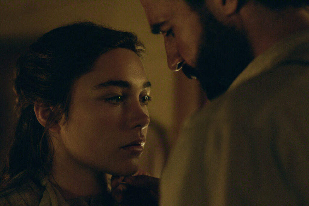 "A young woman and a bearded man stand closely in a dimly lit room, sharing an intense and intimate moment. The scene has a soft, warm glow and a serious mood, suggesting a private, emotional exchange between the two."